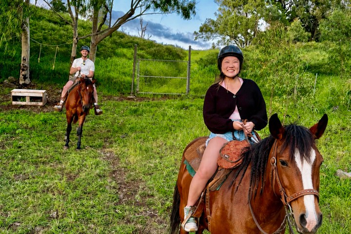 Two people horseback riding on a grassy trail with trees.