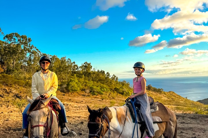 Two people on horses in a scenic landscape with a bright blue sky and white clouds.