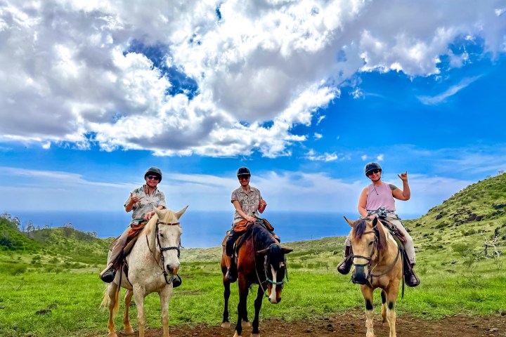 Three people on horses in a scenic landscape with blue sky and clouds.