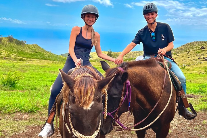Two people horseback riding in a grassy landscape with a blue sky.