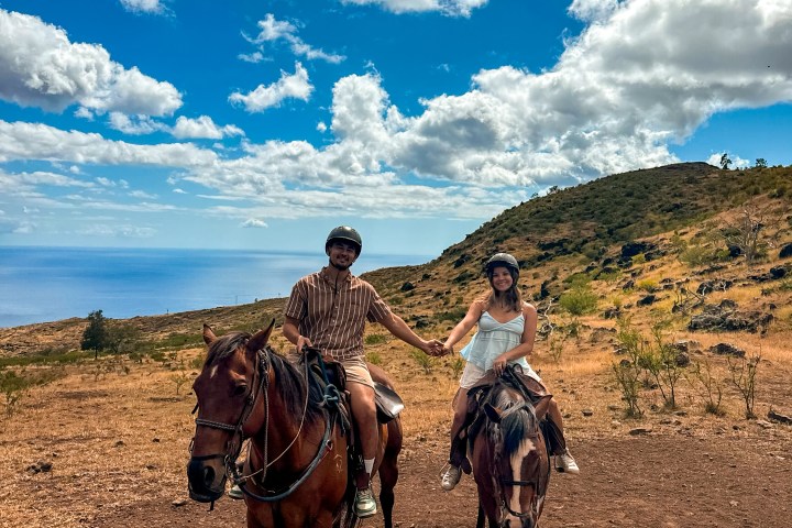 Two people holding hands while horseback riding on a hill with blue sky and clouds.