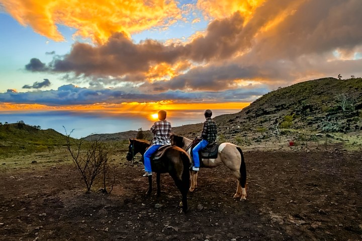 Two people riding horses at sunset with dramatic orange clouds.