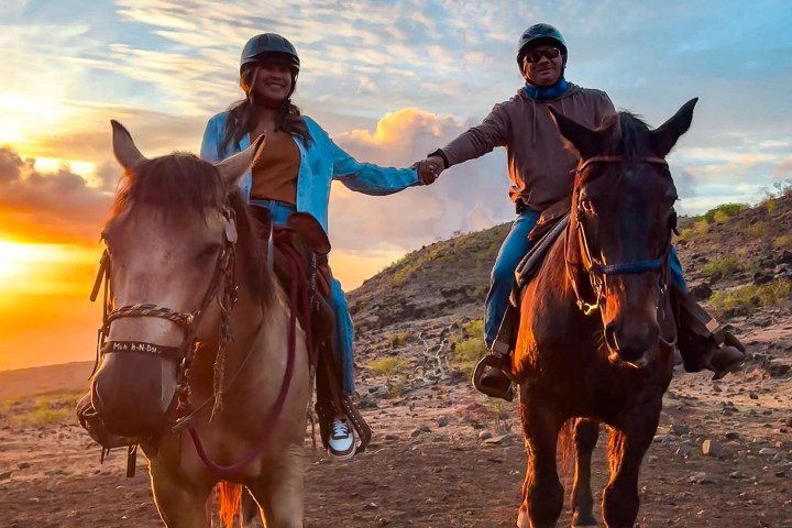 Two people horseback riding at sunset, holding hands, under a partly cloudy sky.