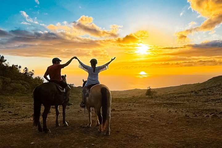Two people on horses hold hands under a vibrant sunset sky.