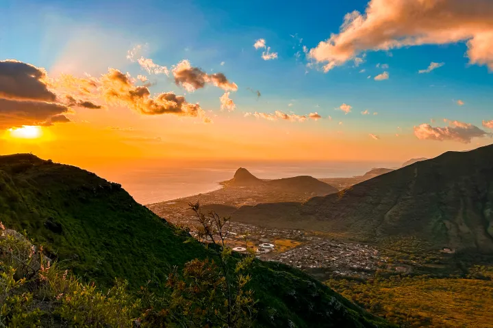Scenic sunset over a lush valley with mountains, ocean, and a glowing sky with scattered clouds.