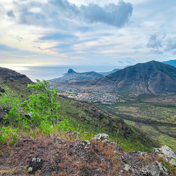 Mountainous landscape with green vegetation and distant ocean under a cloudy sky.