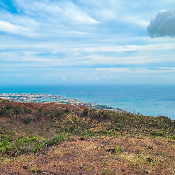 Coastal landscape with hills, scattered buildings, and a cloudy blue sky.