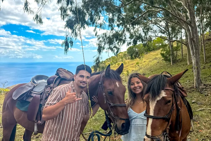 Two people with two horses in a grassy area with trees and a view of the ocean.