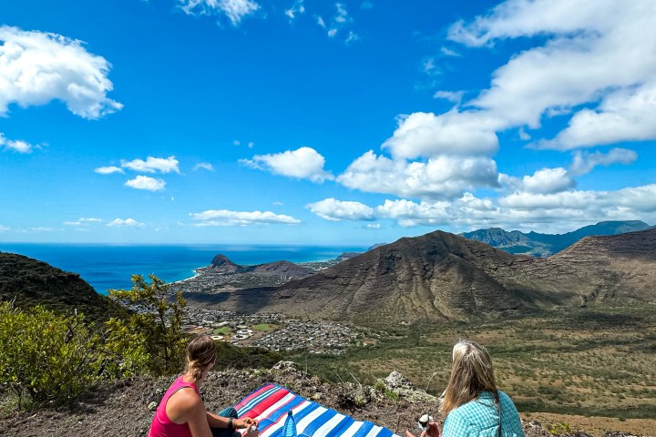 Two people picnicking on a hilltop with a scenic view of mountains and ocean under a blue sky.