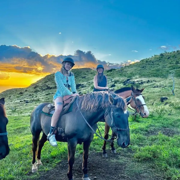 Two people riding horses on a green hillside at sunset with a blue sky.
