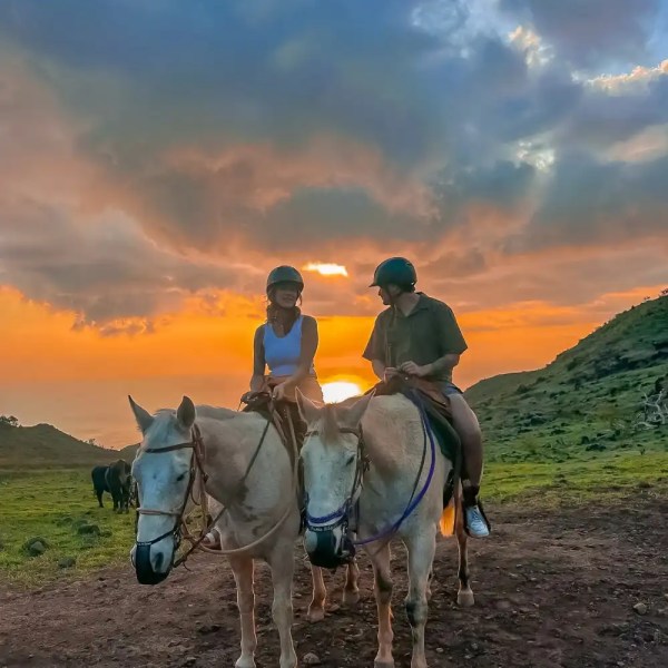 Two people riding horses at sunset with a dramatic sky background.