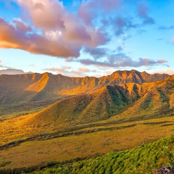 Sunlit mountain range with green slopes under a colorful sky with clouds.