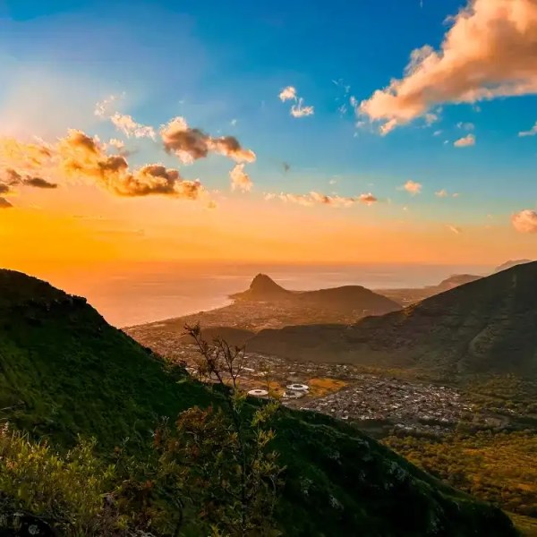 Sunset over coastal valley with hills, green vegetation, and scattered clouds.
