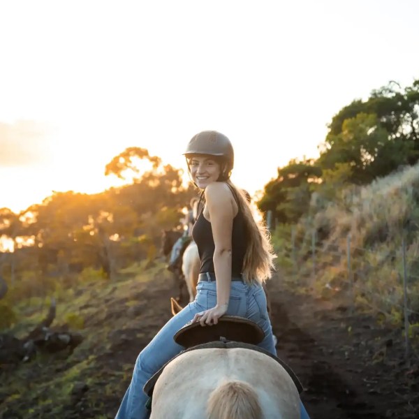 Woman riding horse on a trail during sunset, wearing a helmet and smiling.