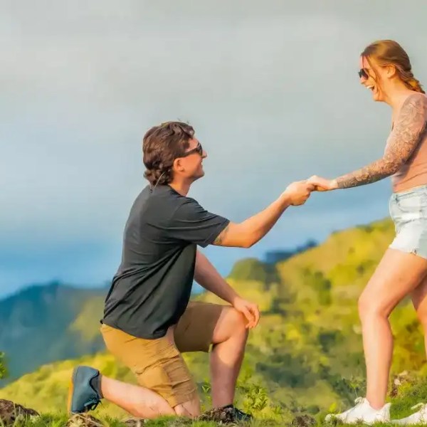 Man kneels proposing to woman, both smiling, on a grassy hill with mountains in background.