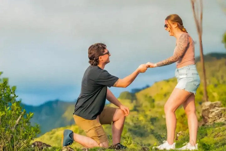 Man kneels proposing to woman, both smiling, on a grassy hill with mountains in background.